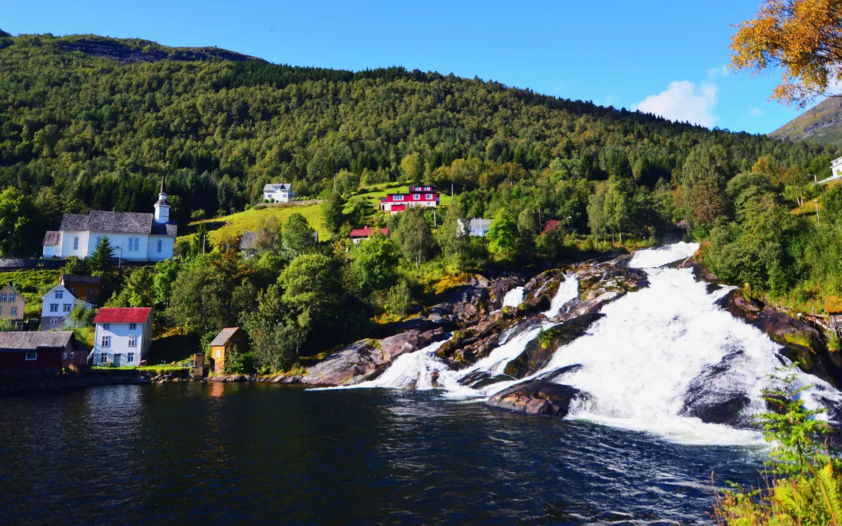 Wasserfall in Hellesylt, umgeben von grünen Hügeln und Häusern, bei sonnigem Wetter.