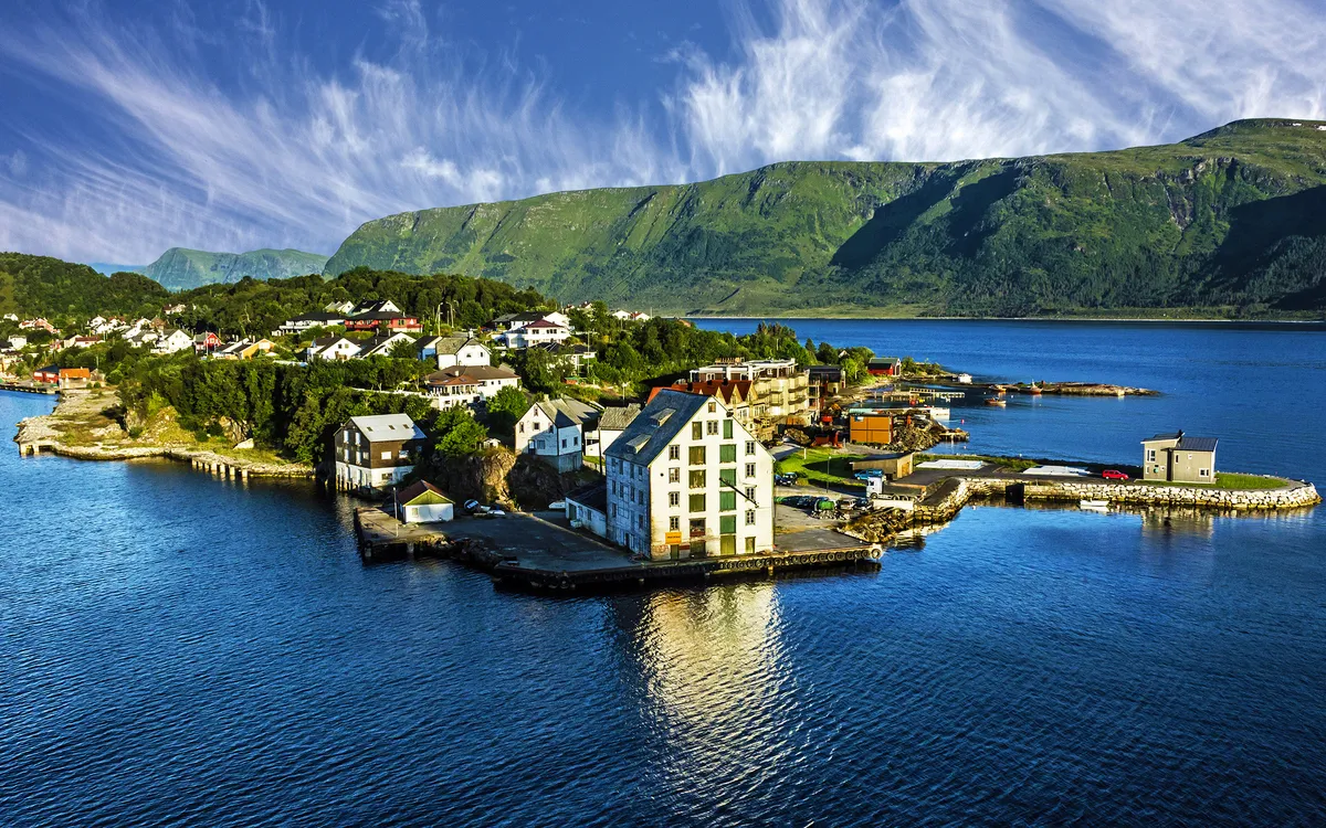 Küste mit kleinen Gebäuden auf einer Insel, umgeben von blauem Wasser und Bergen im Hintergrund.