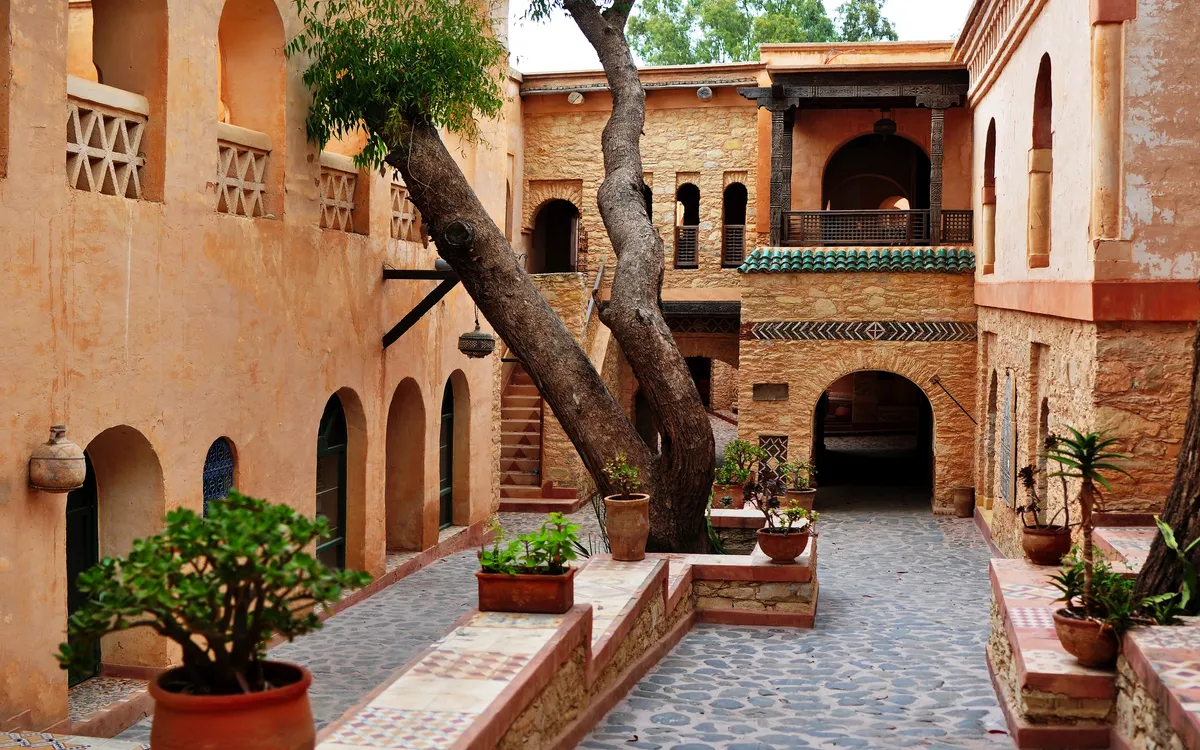 Innenhof mit Baum, Topfpflanzen und antikem Gebäude in warmen Farbtönen.