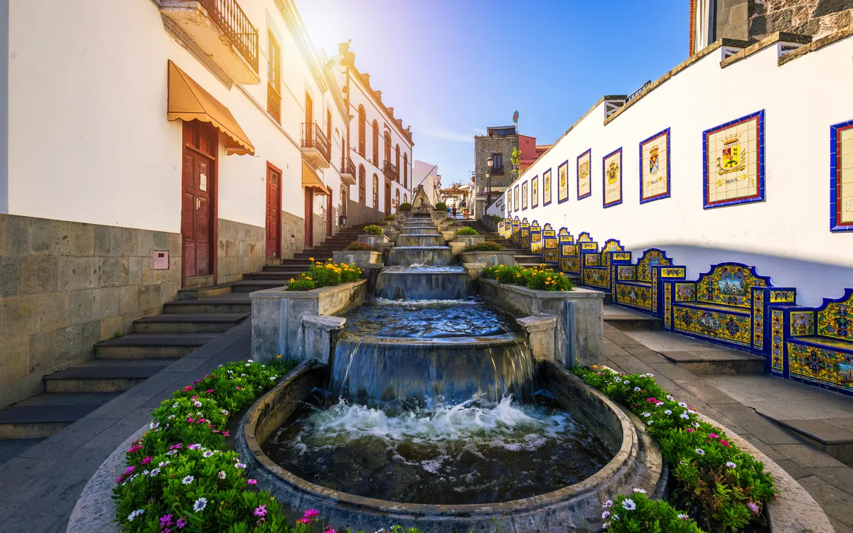 Kaskadierende Springbrunnen in sonniger Straße mit verzierten Wänden und Blumen.