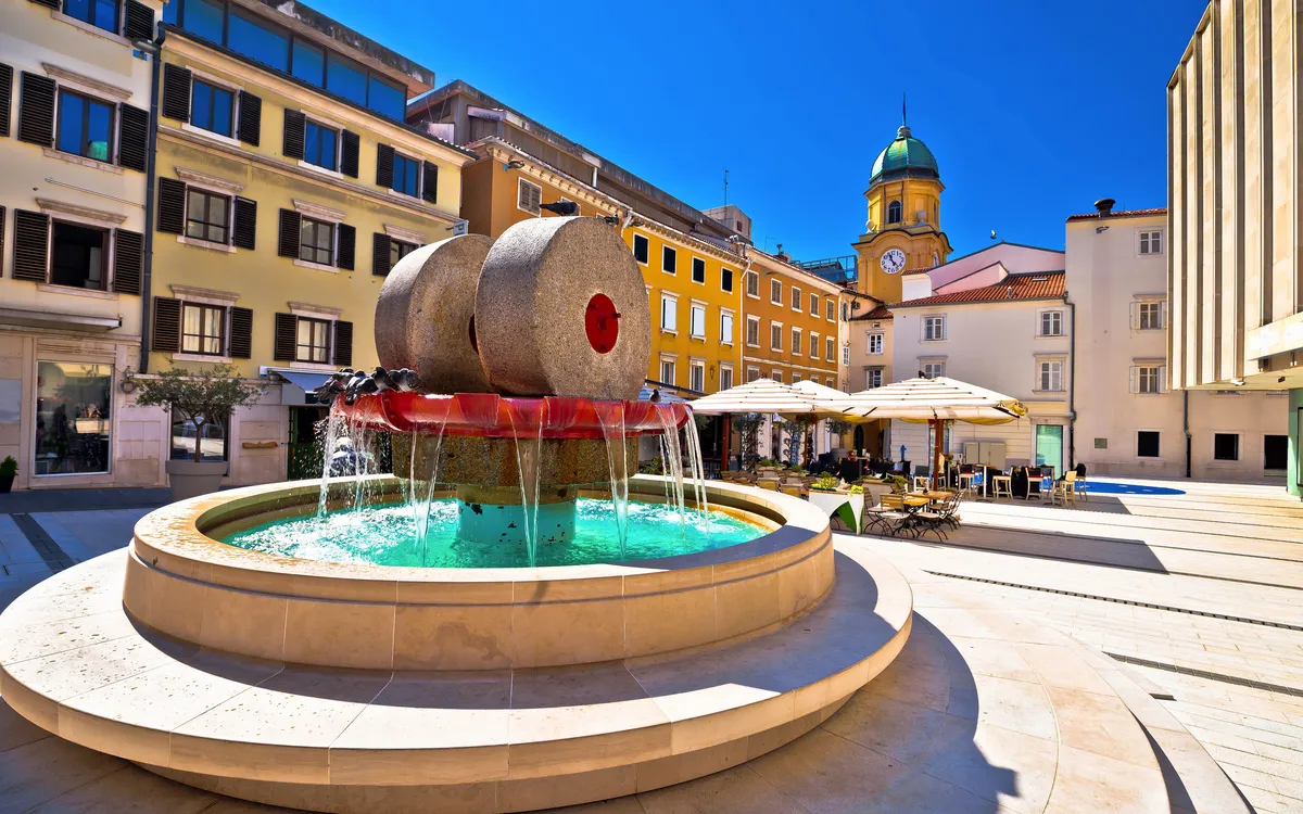 Brunnen auf einem Platz mit umliegenden historischen Gebäuden und blauem Himmel.