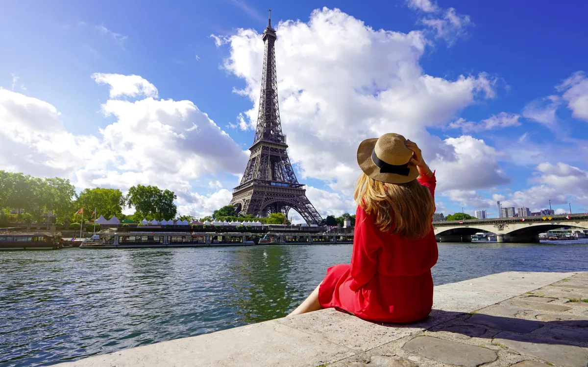 Reisende in rotem Kleid und Hut sitzt am Ufer der Seine in Paris und blickt auf den Eiffelturm.