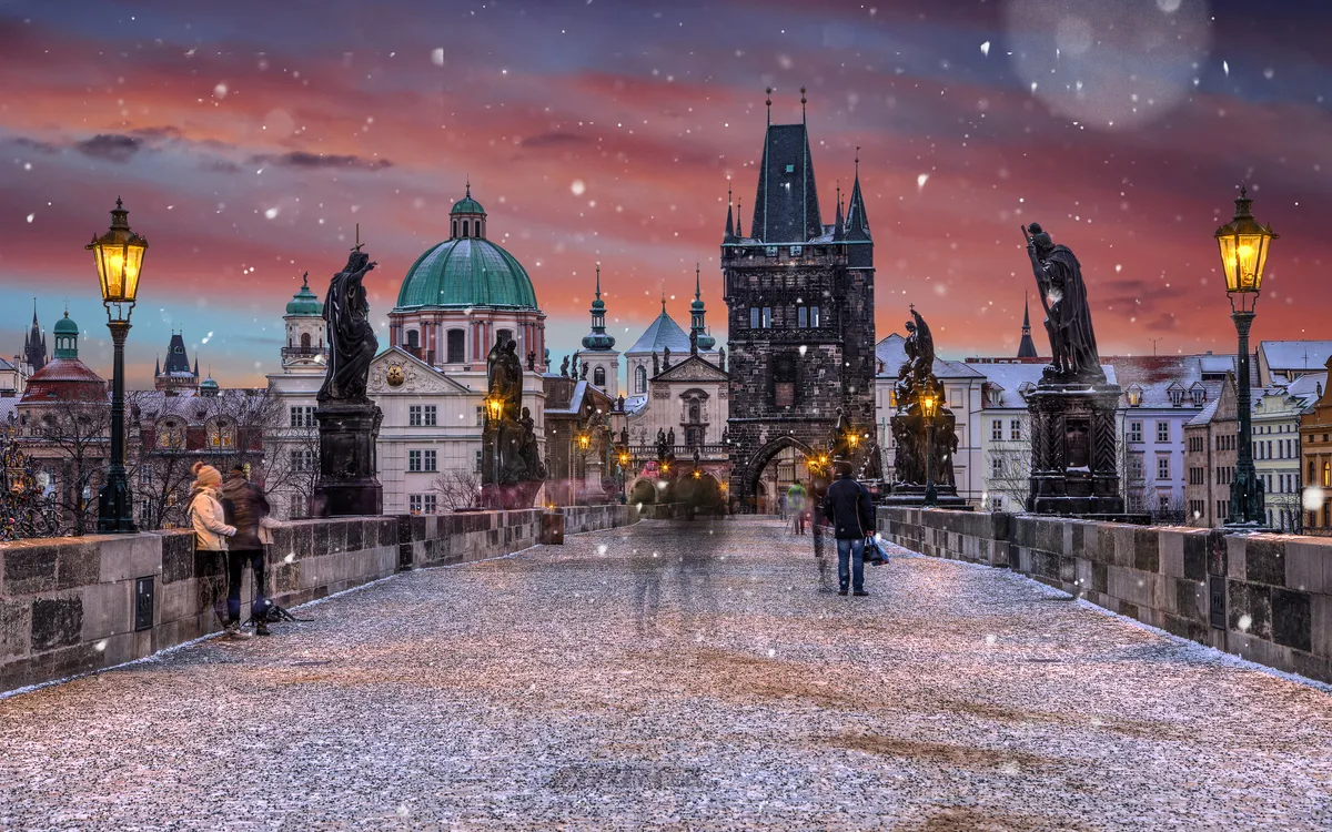 Menschen spazieren bei Schneefall über die Karlsbrücke in Prag bei Sonnenuntergang.