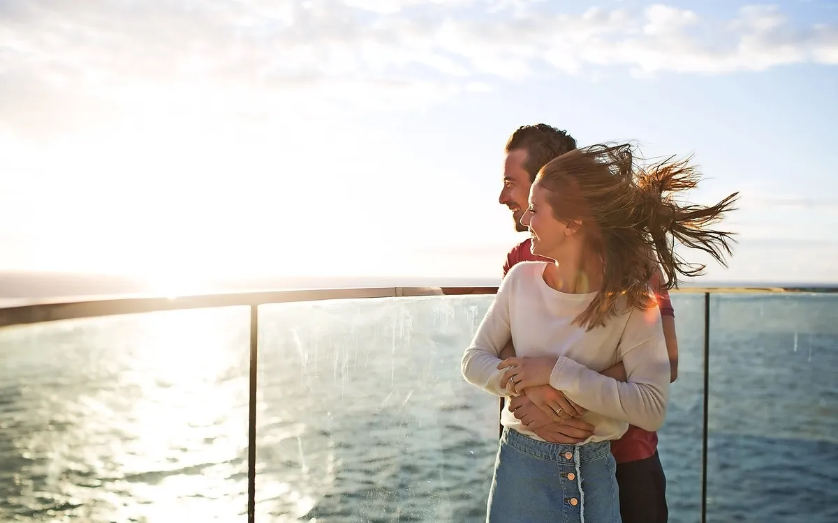 Pärchen umarmt sich lachend auf einem Skywalk über dem Meer bei Sonnenschein.