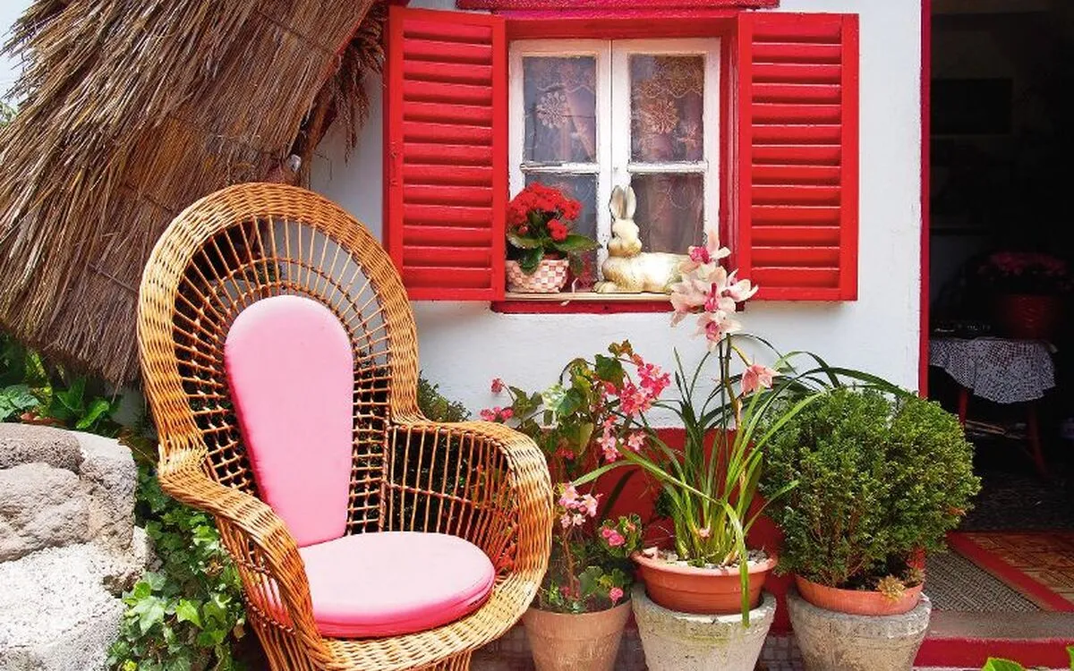 Traditionelles Santana-Häuschen mit roten Fensterläden und Korbstuhl, umgeben von Pflanzen.