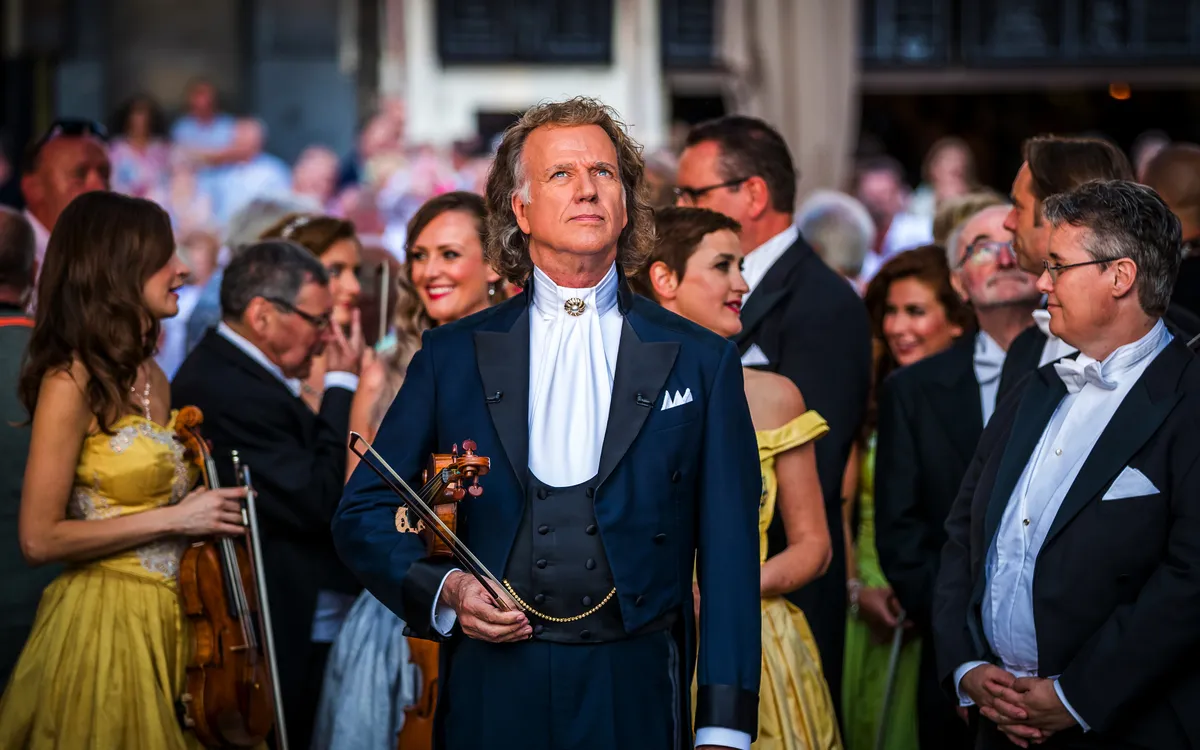 Orchester dirigiert von Mann in dunklem Anzug bei Open-Air-Konzert mit Zuschauern im Hintergrund.