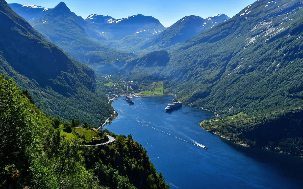 Blick auf einen Fjord mit Bergen und Kreuzfahrtschiffen bei sonnigem Wetter.