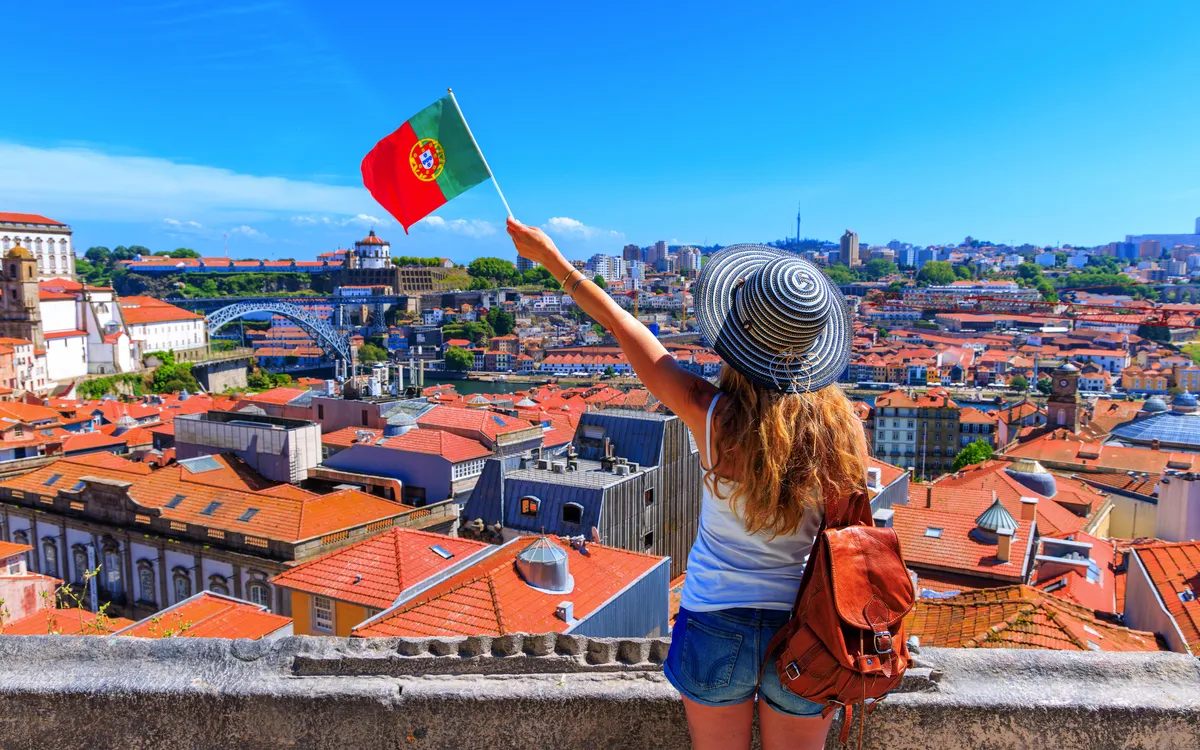 Person mit Hut schwenkt portugiesische Flagge über Stadt mit roten Dächern.