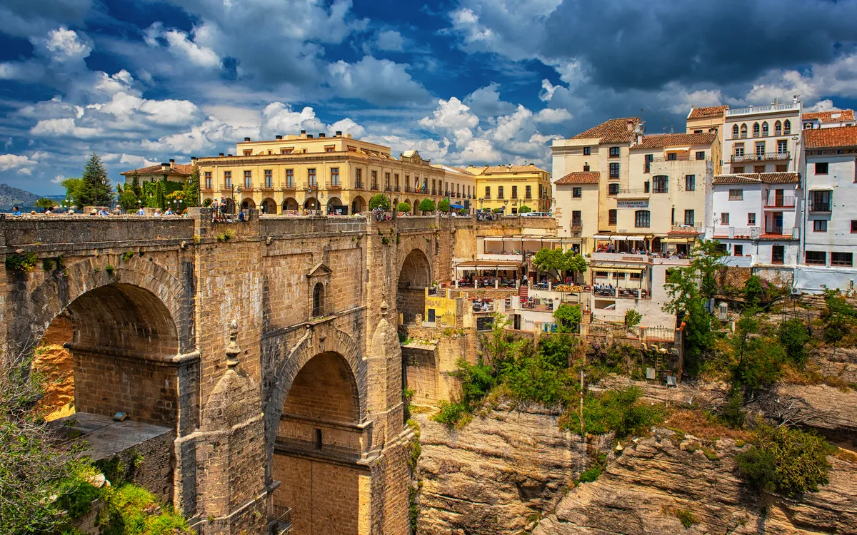 Historische Steinbrücke in Ronda, umgeben von Gebäuden und bewölktem Himmel.