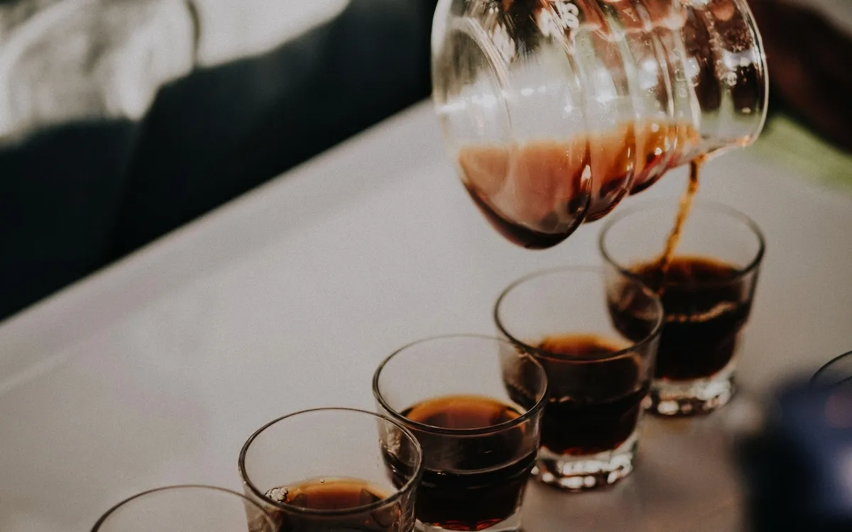Hand gießt Kaffee in mehrere kleine Gläser auf einem Tisch.