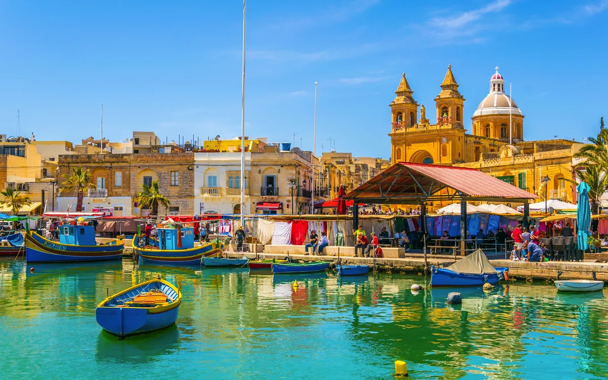 Bunte Fischerboote im Hafen vor historischen Gebäuden und einem Markt in Malta.
