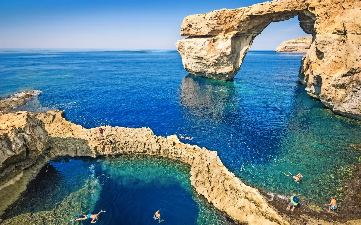 Felsformation im Meer mit natürlichem Bogen, klare blaue Gewässer, Schwimmer im Vordergrund.