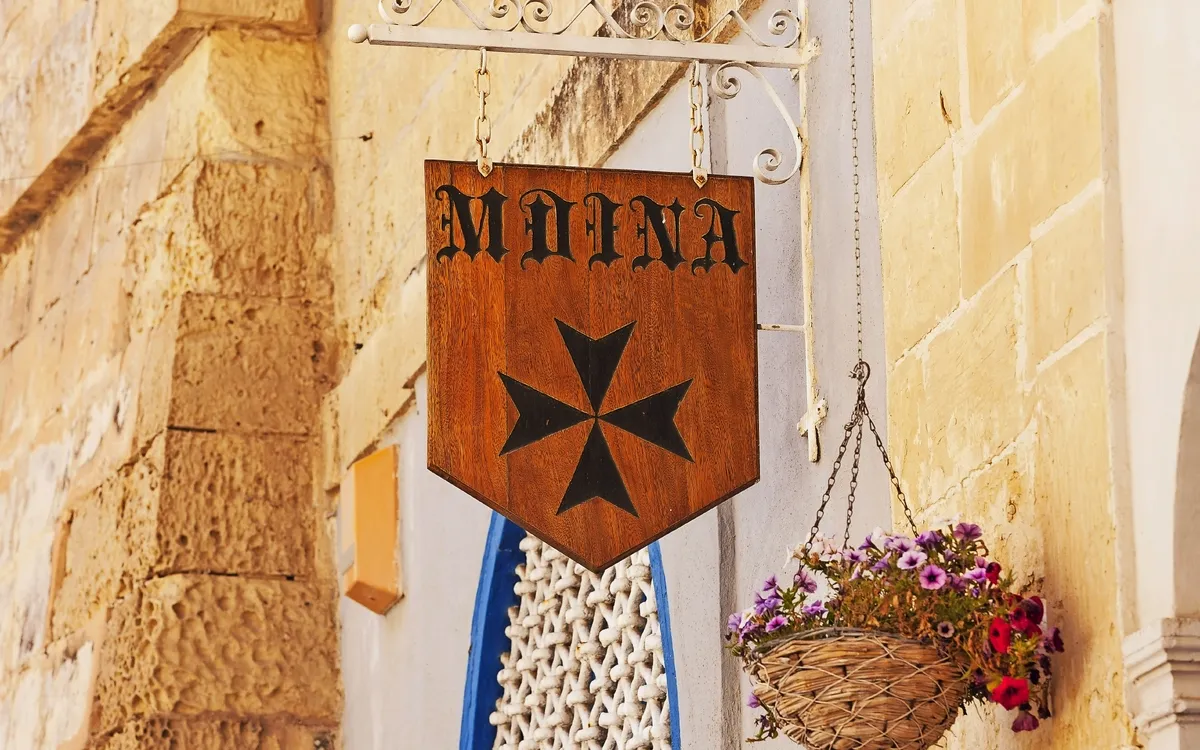 Holzschild mit Malteserkreuz und Schriftzug Mdina, daneben Blumenampel an einer Steinwand.