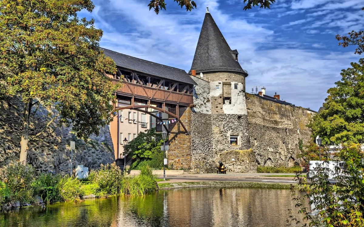 Historischer Turm und Mauer mit Wassergraben und Bäumen im Vordergrund.